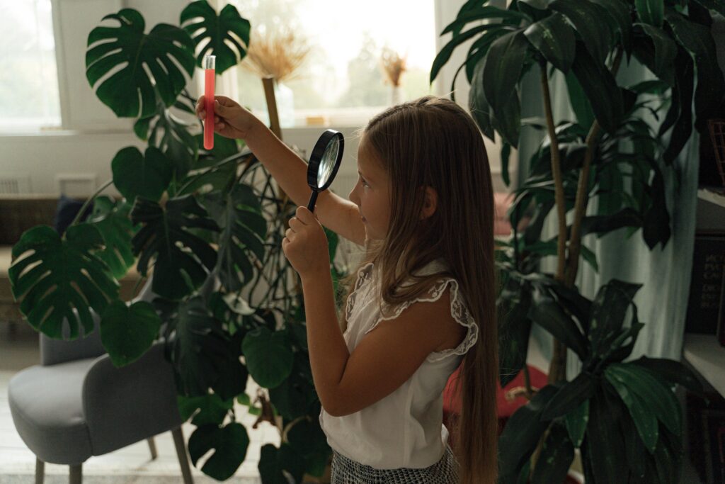 A young girl examines plants with a magnifying glass and test tube indoors.